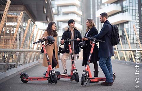 Zwei Frauen und zwei Männer stehen mit voi E-Scootern in einer urbanen Umgebung in einer Gruppe zusammen.