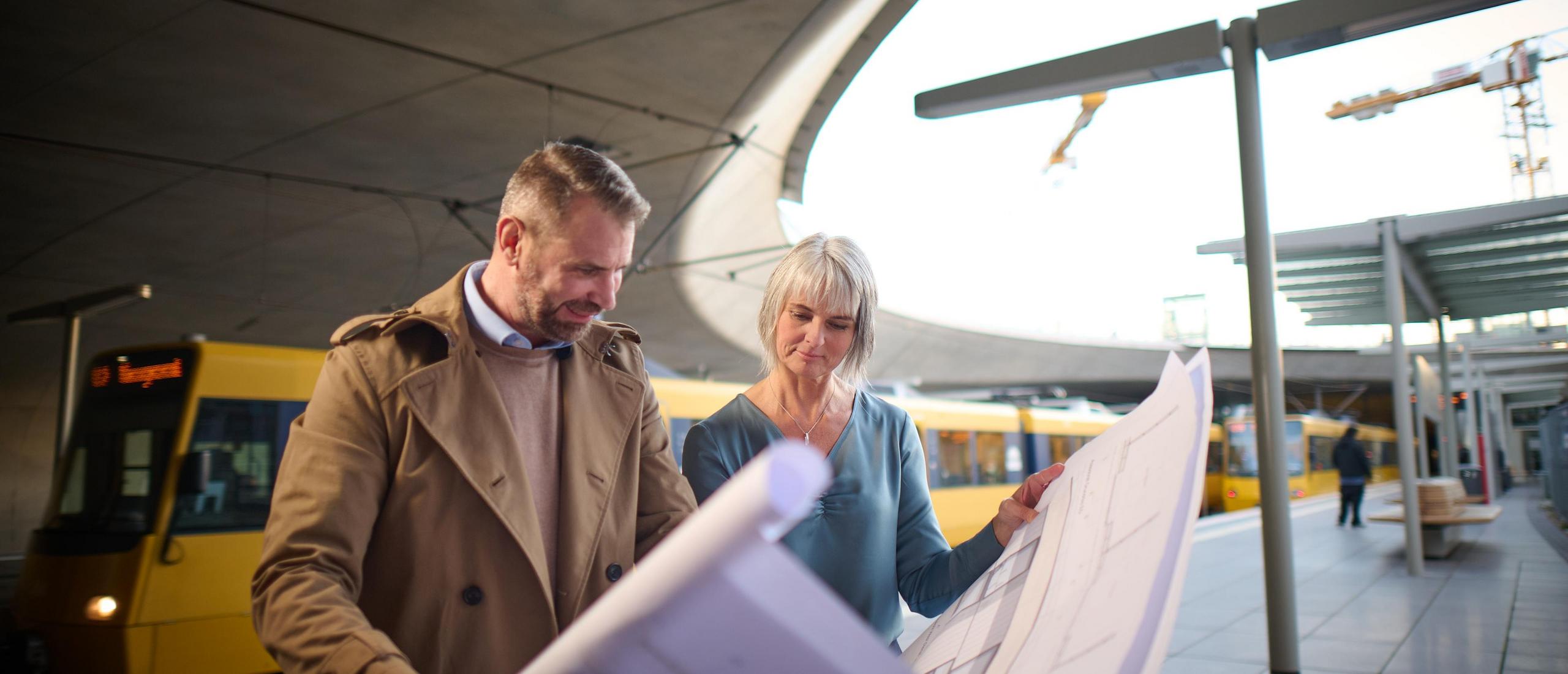 Zwei Architekten schauen sich auf einem Bahnsteig einen Skizzenplan an.