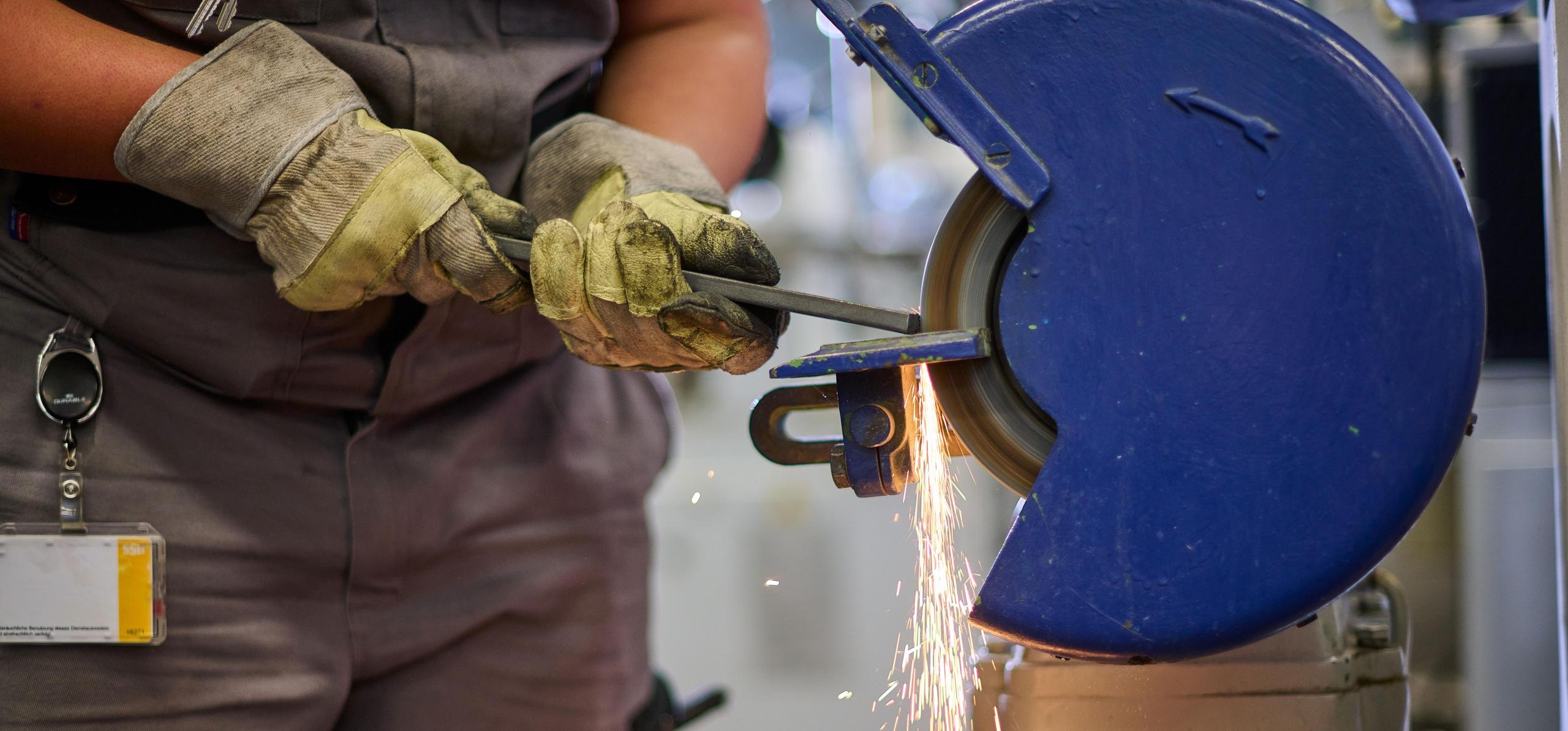Zwei Hände halten eine Eisenstange und schleifen es an einer elektrischen Schleifanlage.