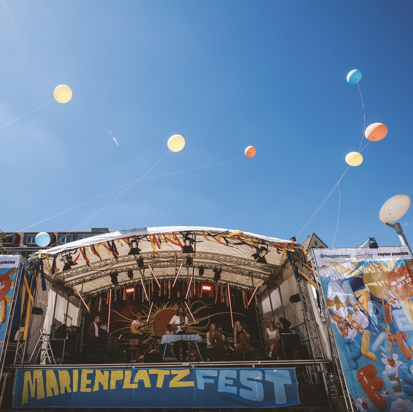 Auf der Bühne spielt eine Band am Marienplatzfest und Luftballons ragen in den Himmel.