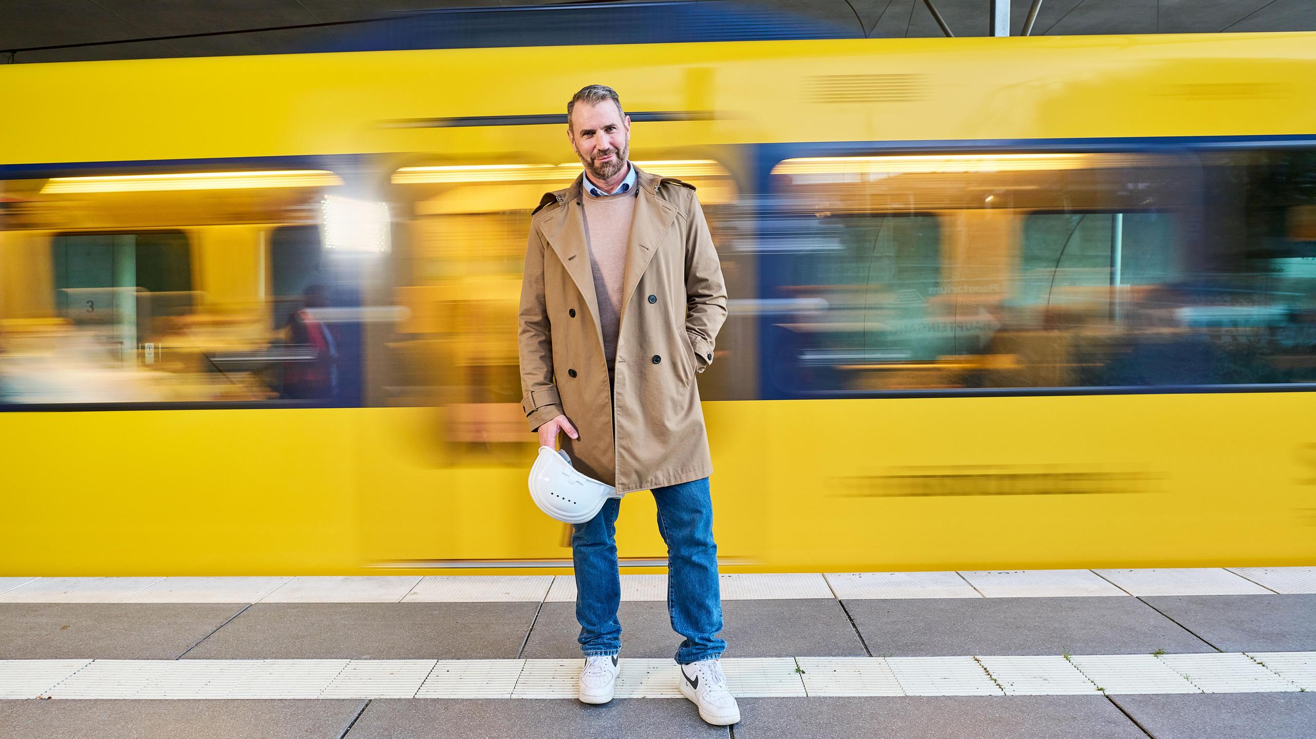 Ein Mann mittleren Alters hält einen Bauhelm in der Hand, während hinter ihm eine Stadtbahn fährt.