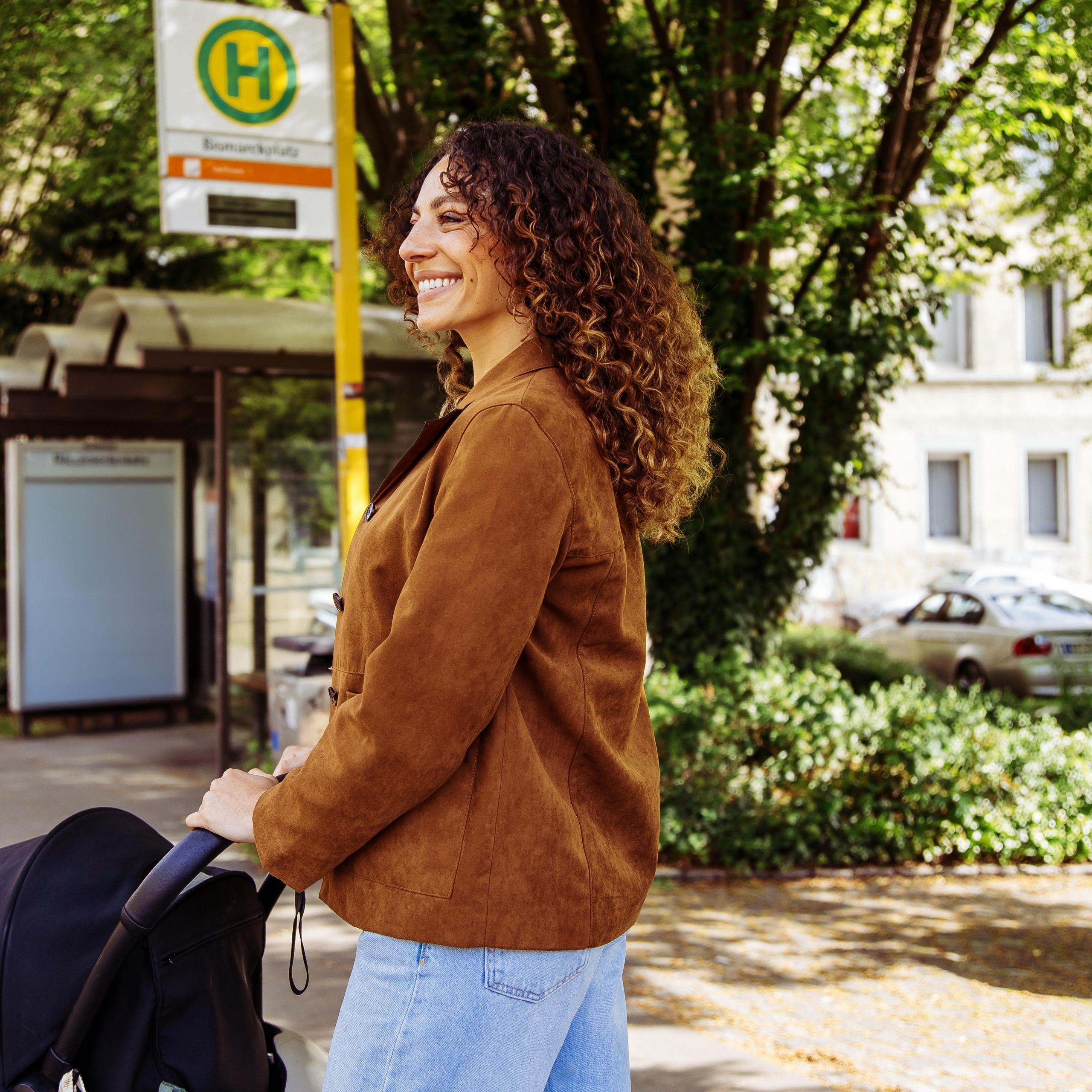 Junge Frau, die mit Kinderwagen an der Bushaltestelle steht und lächelnd auf den Bus wartet.