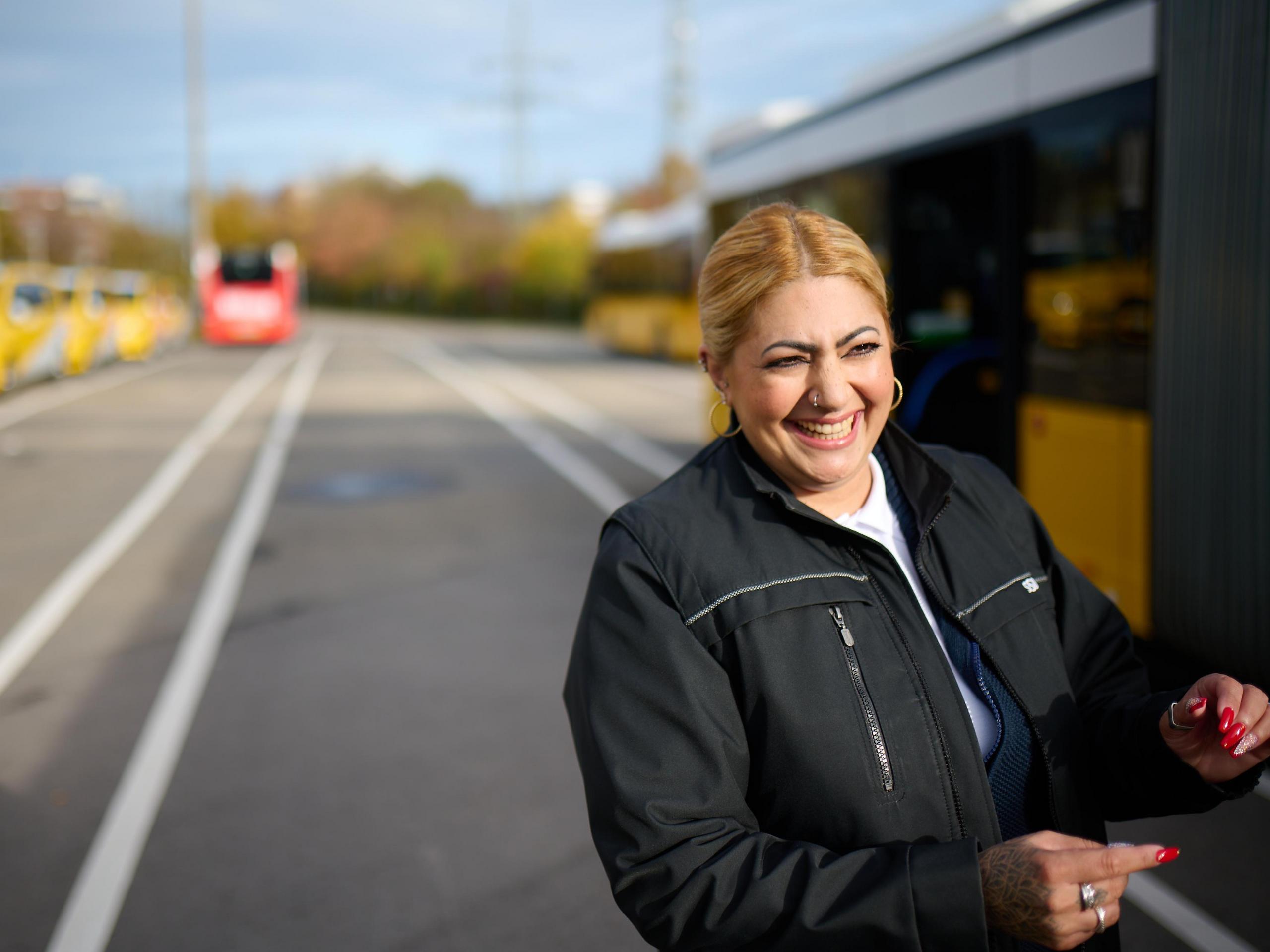 Eine Busfahrerin steht neben einem Bus und lacht.