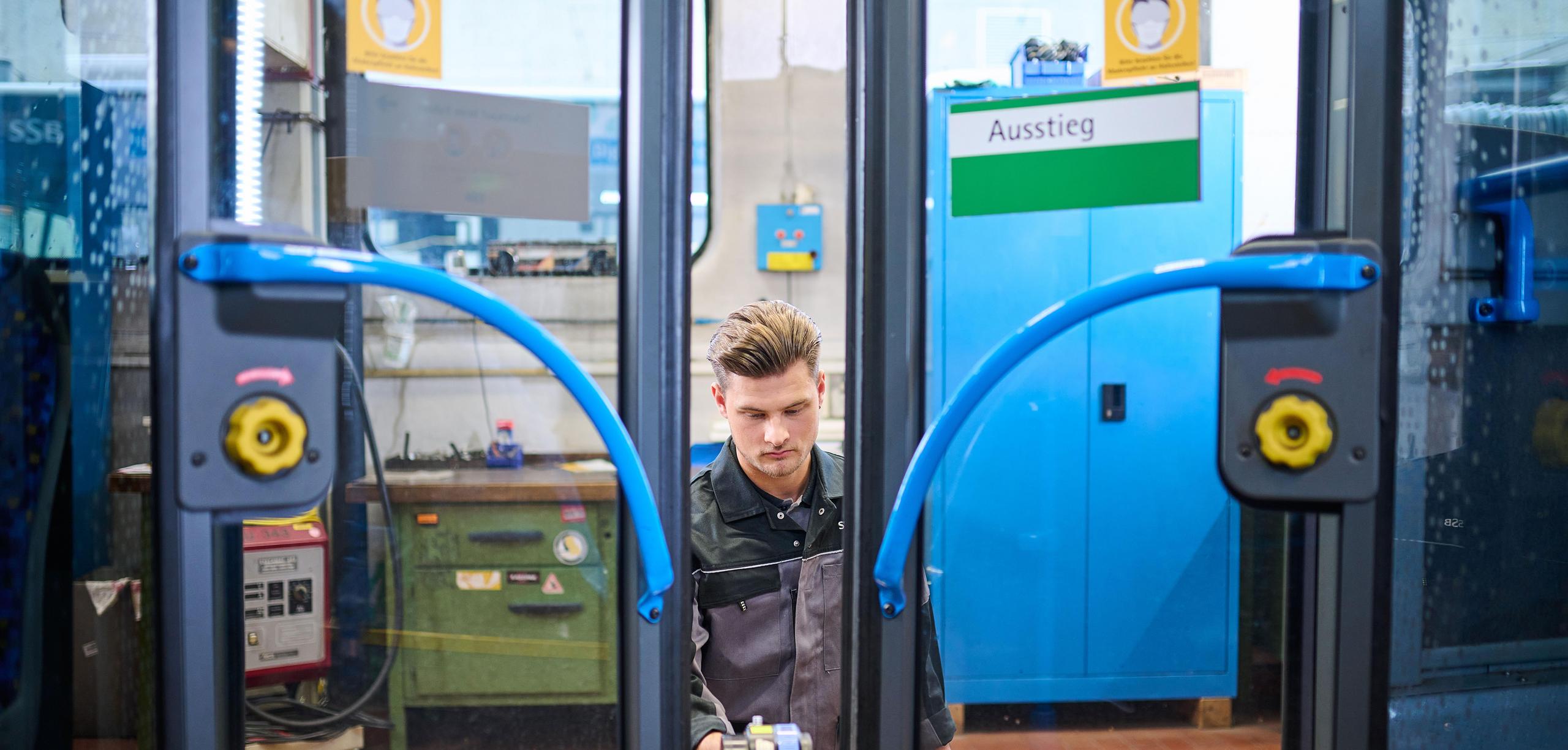 Ein Auszubildender in Werkstattkleidung prüft die Türen eines Busses.