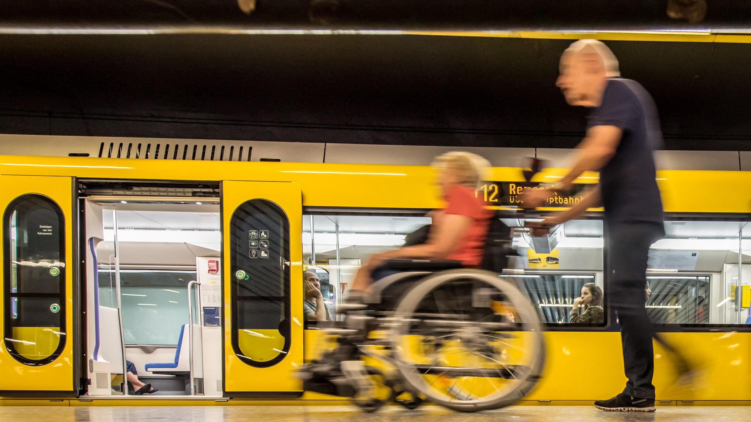 Ein Mann schiebt eine ältere Dame im Rollstuhl entlang einer Stadtbahn.