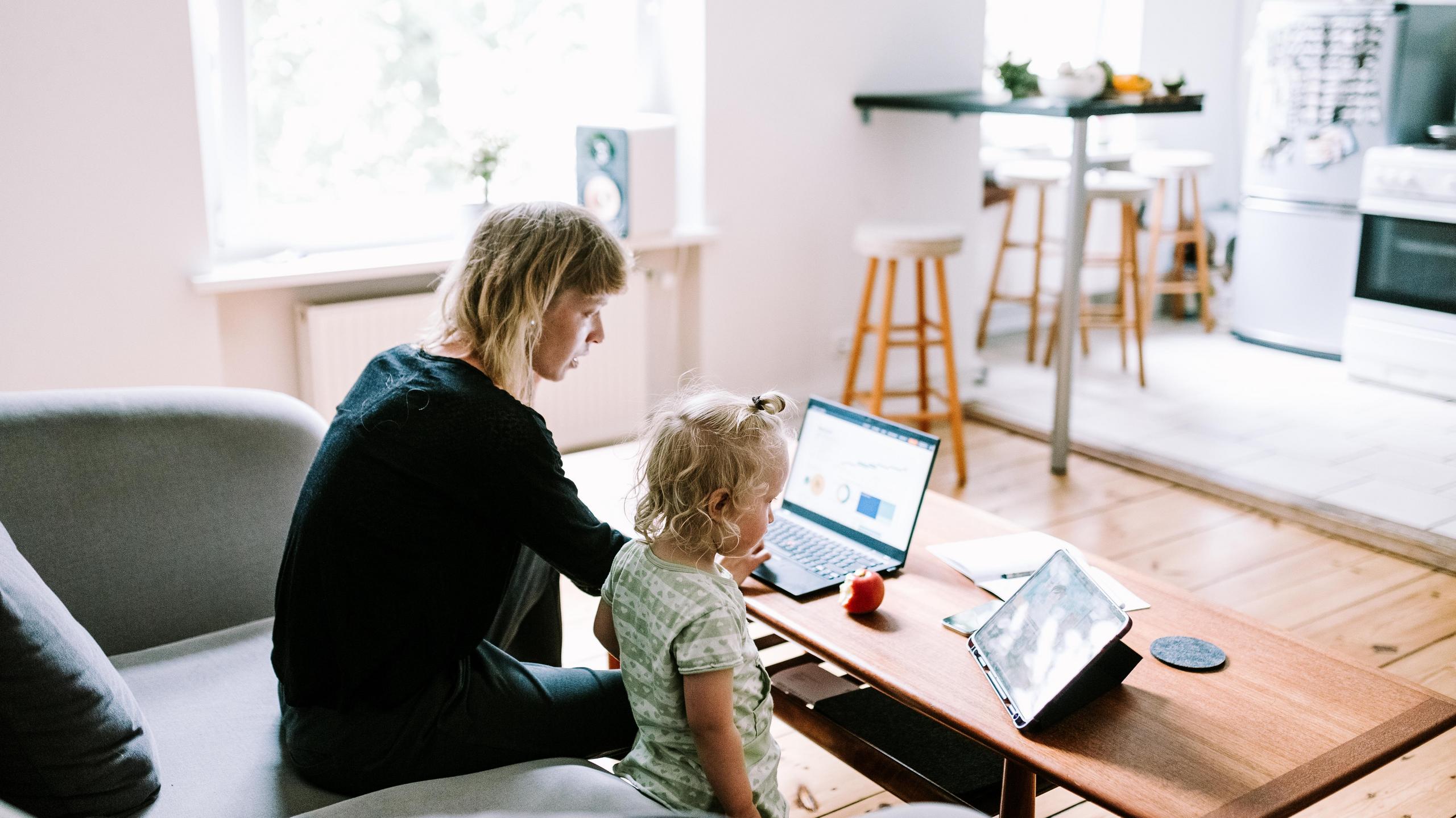 Eine Mutter sitzt auf einem Sofa und schaut auf ihren Laptop. Daneben steht ein kleines Kind.