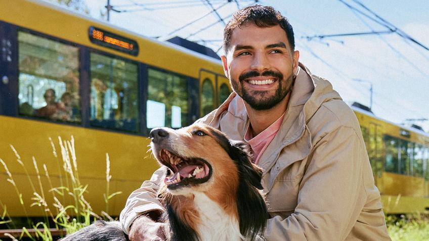 Ein junger Mann mit einem Hund vor einer vorbeifahrenden Stadtbahn.