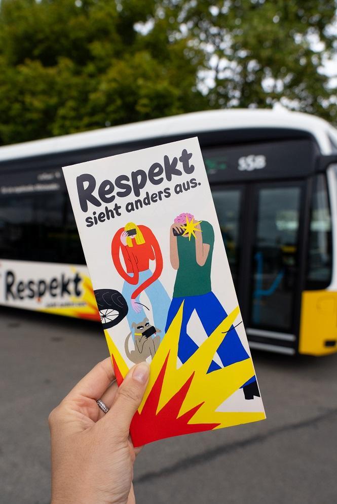 Ein Flyer der Respekt-Kampagne wird in die Kamera gehalten im Hintergrund ist der Bus zur Kampagne zu sehen.